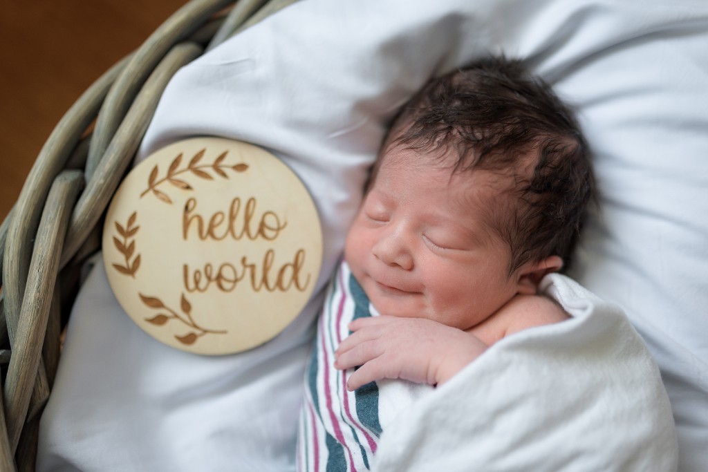Newborn in a basket with a hello world wooden sign